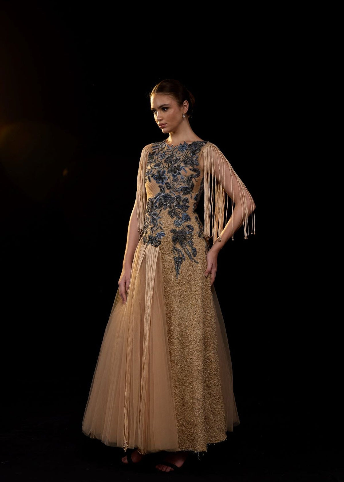 A nude toned gown adorned with intricate blue floral embroidery and dramatic fringe sleeves.