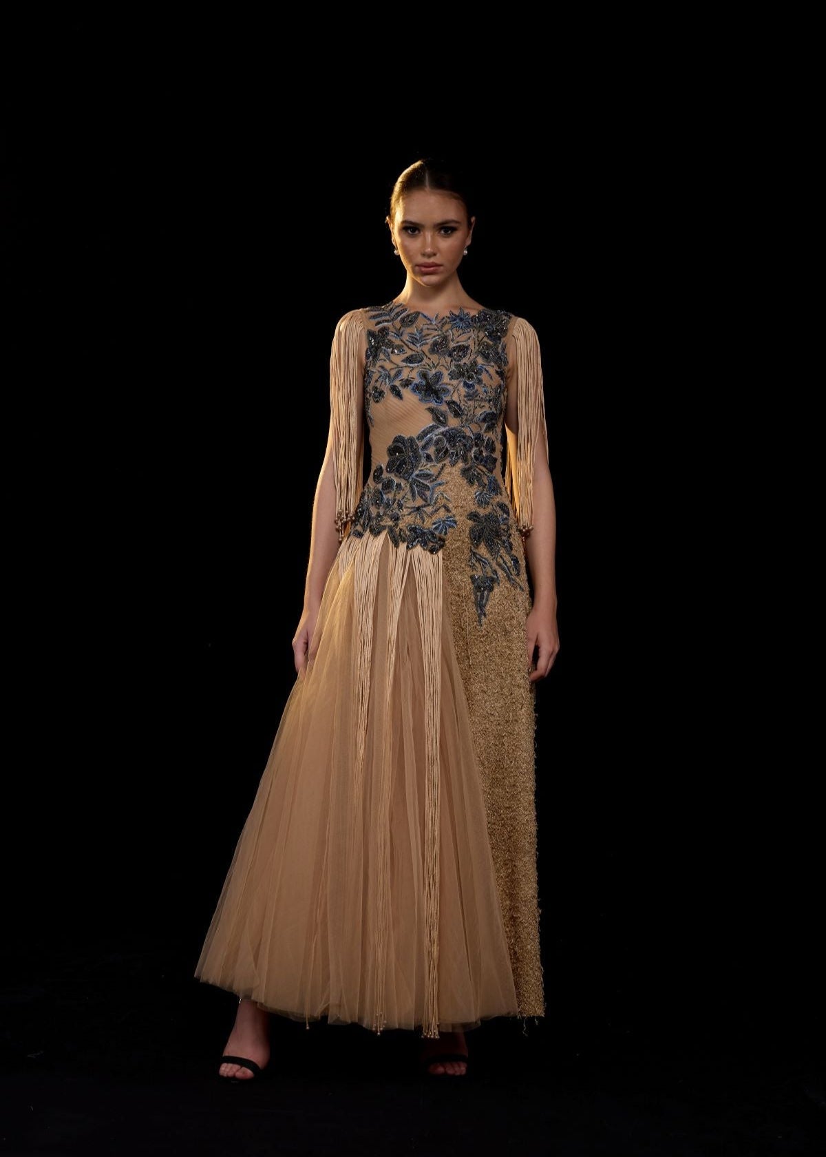A nude toned gown adorned with intricate blue floral embroidery and dramatic fringe sleeves.