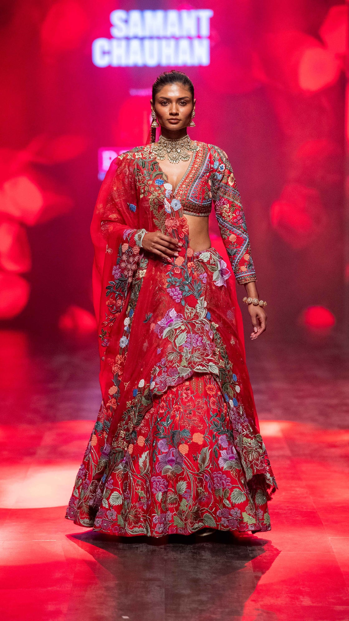 Model wearing a vibrant red and multicolor traditional outfit on a stage with 'Samantha Chauhan' branding.
