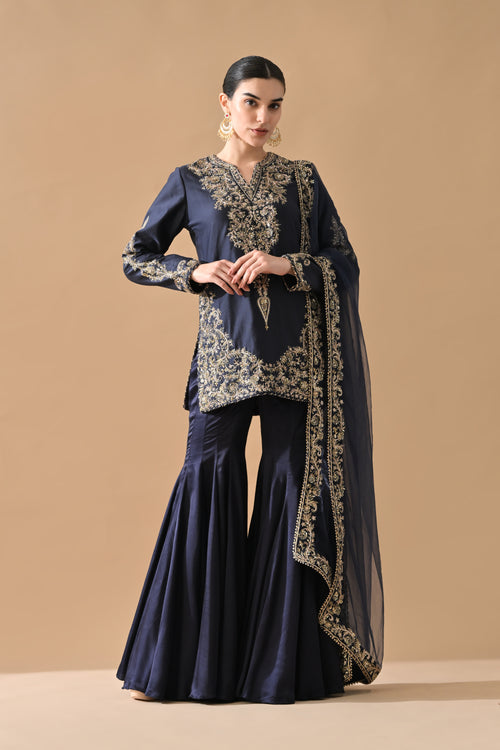 Woman wearing a navy blue traditional outfit with gold embroidery on a beige background