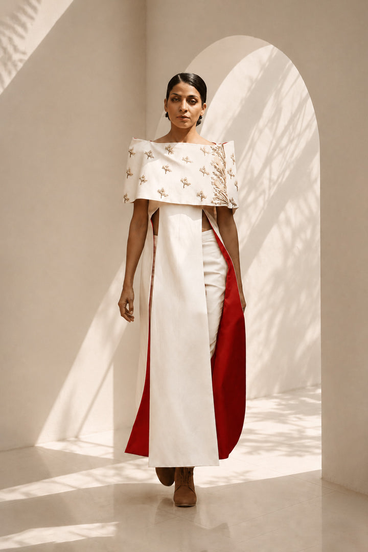 Ivory striking architectural ensemble featuring a sculptural cape overlay, dramatic side panels, and contrasting crimson accents that redefine contemporary elegance.