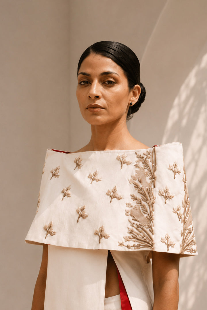 Ivory striking architectural ensemble featuring a sculptural cape overlay, dramatic side panels, and contrasting crimson accents that redefine contemporary elegance.