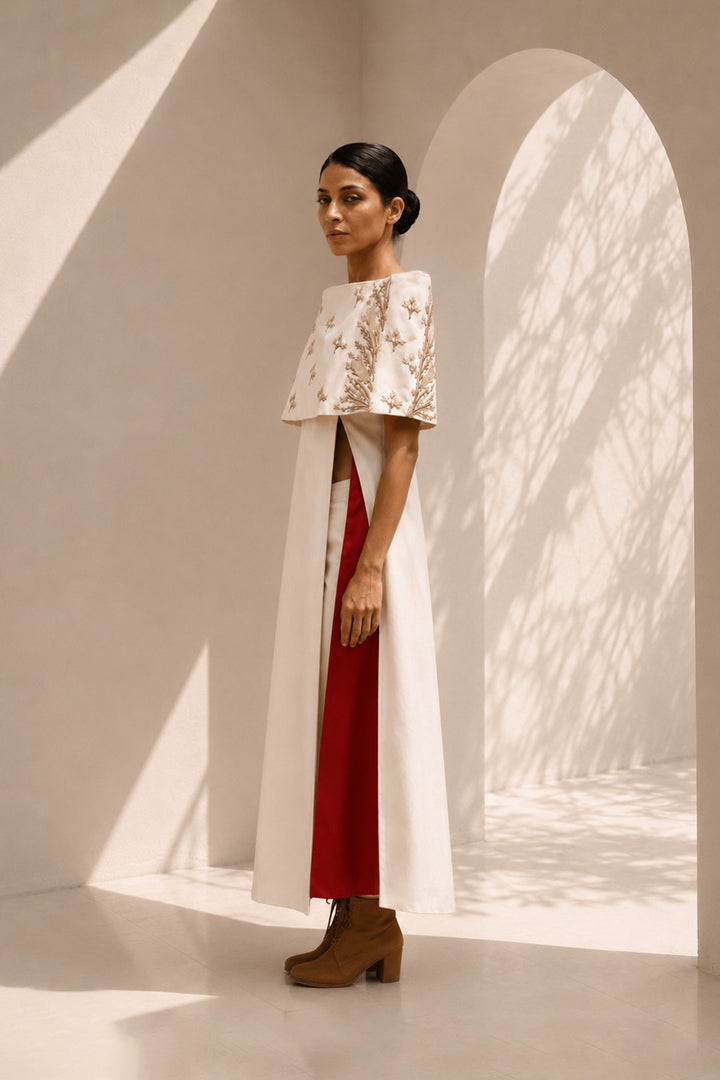 Ivory striking architectural ensemble featuring a sculptural cape overlay, dramatic side panels, and contrasting crimson accents that redefine contemporary elegance.