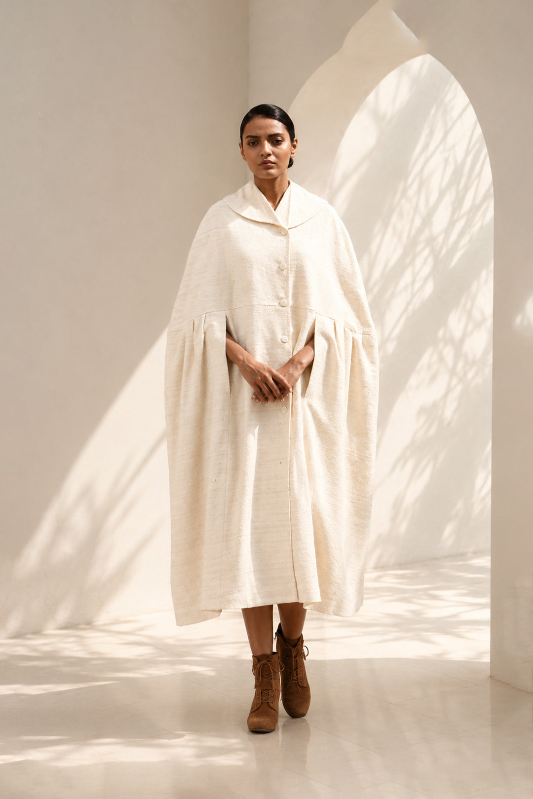 Ivory cape dress crafted in textured fabric, featuring fluid volume, soft pleating, and an effortlessly sophisticated silhouette.