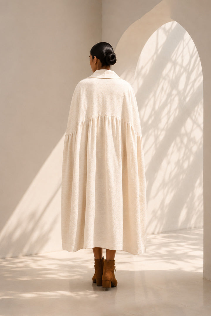 Ivory cape dress crafted in textured fabric, featuring fluid volume, soft pleating, and an effortlessly sophisticated silhouette.
