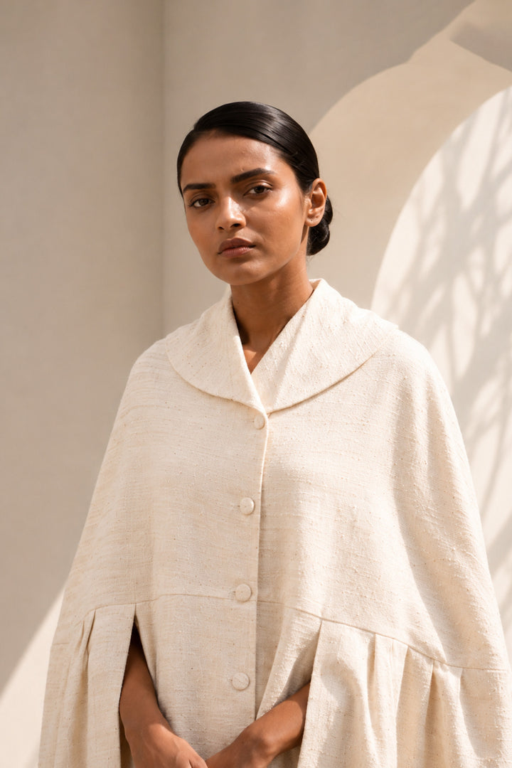 Ivory cape dress crafted in textured fabric, featuring fluid volume, soft pleating, and an effortlessly sophisticated silhouette.