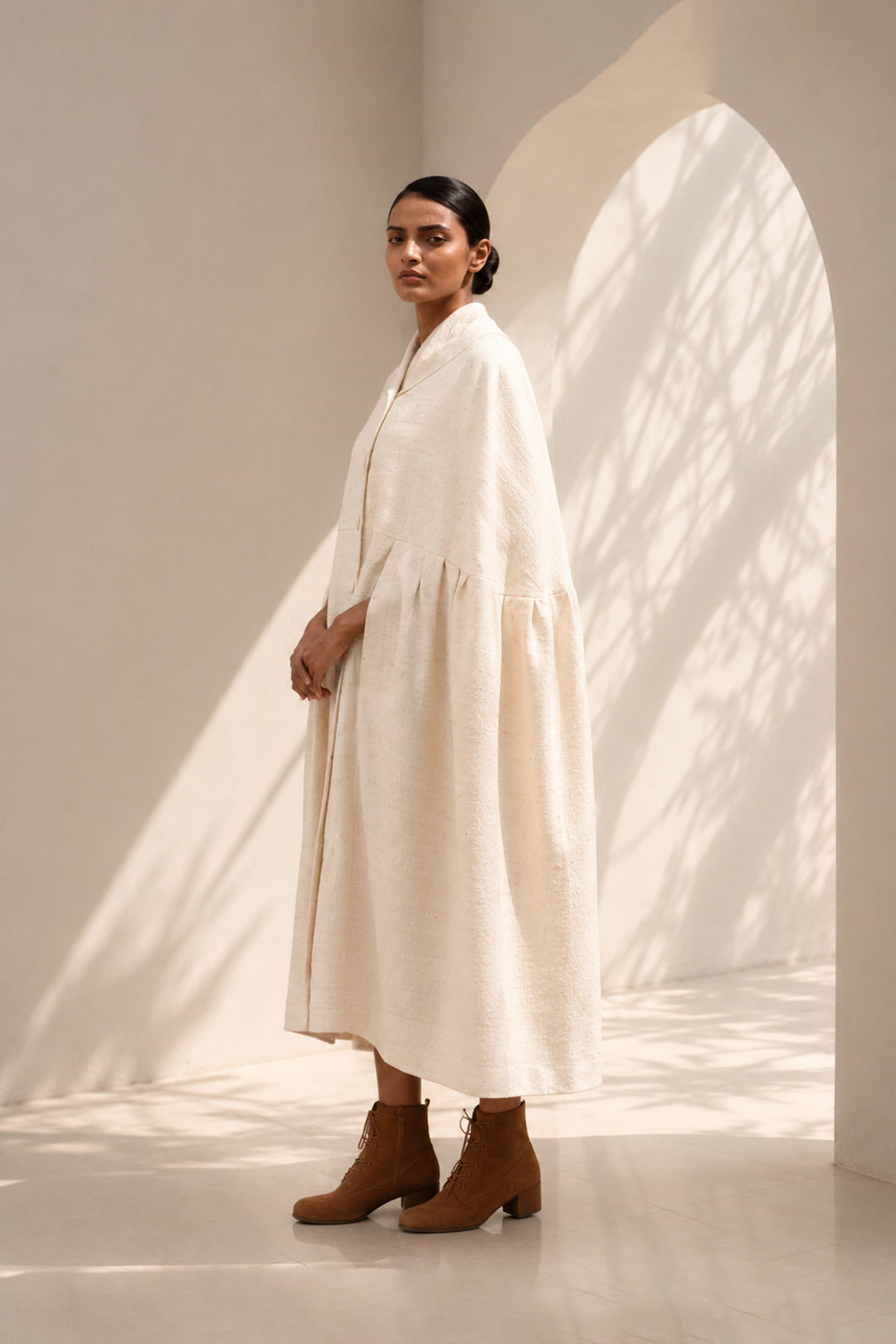 Ivory cape dress crafted in textured fabric, featuring fluid volume, soft pleating, and an effortlessly sophisticated silhouette.