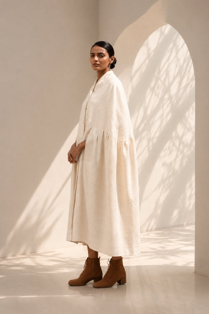 Ivory cape dress crafted in textured fabric, featuring fluid volume, soft pleating, and an effortlessly sophisticated silhouette.