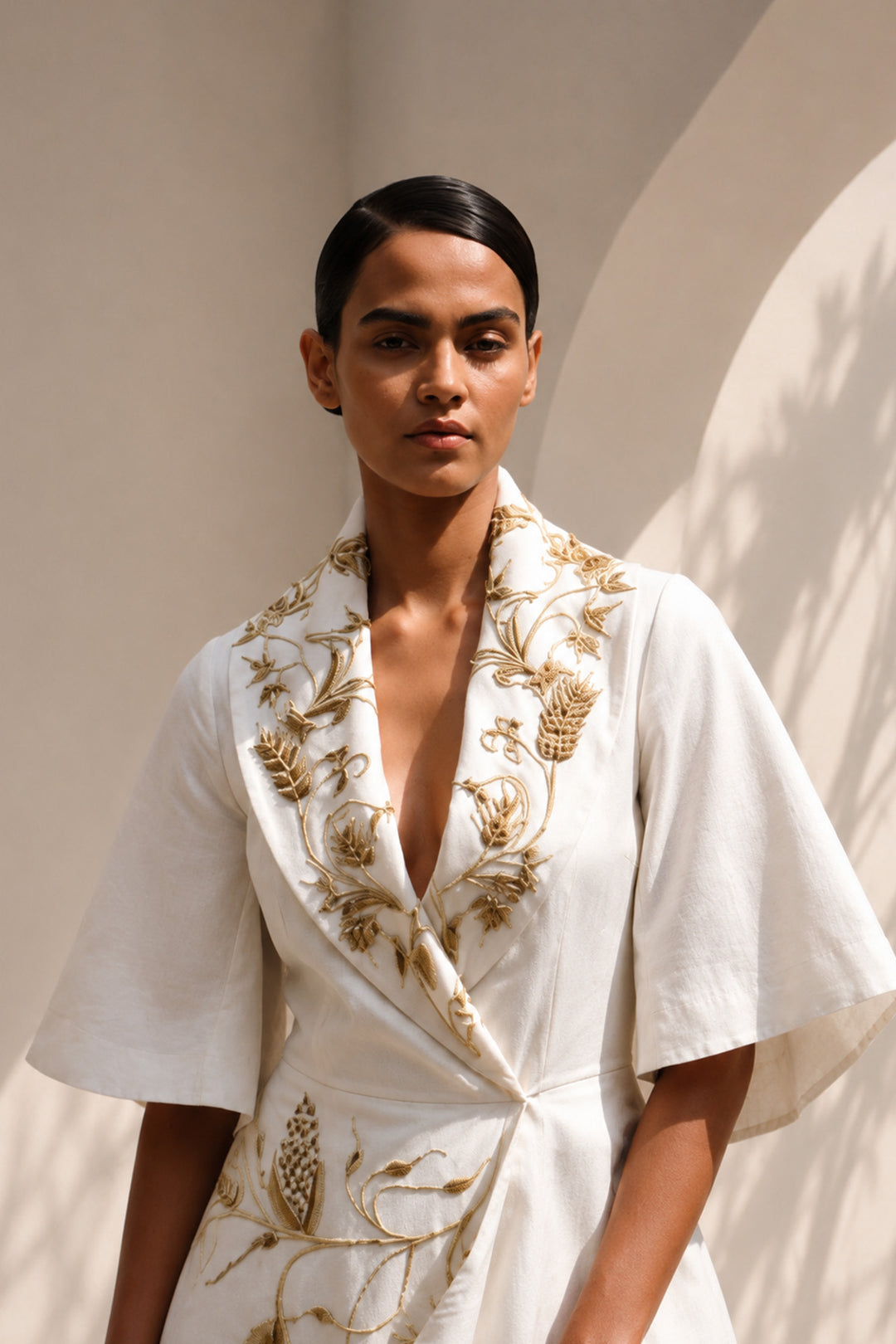 Ivory wrap gown adorned with intricate embroidery, featuring shawl collar and flared sleeves, blending contemporary tailoring with timeless artisanal craftsmanship.