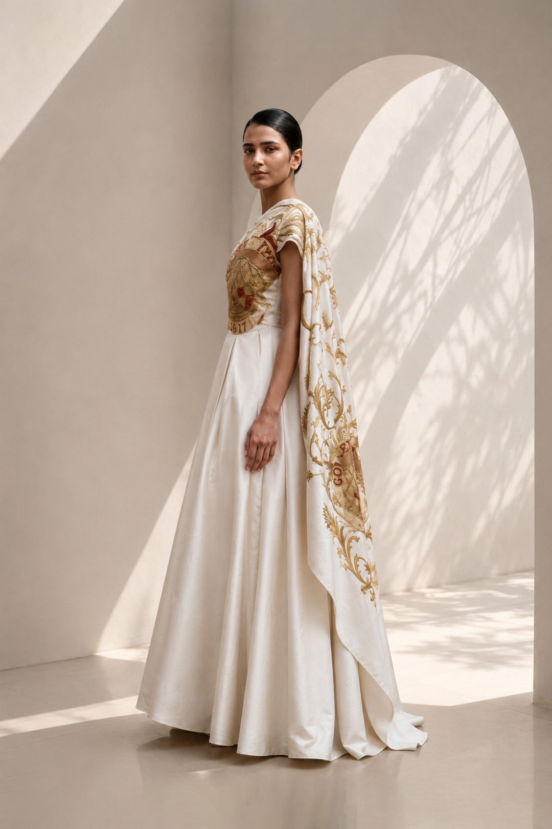 Ivory statement shoulder panel gown featuring gold and crimson artwork and sculptural draping.