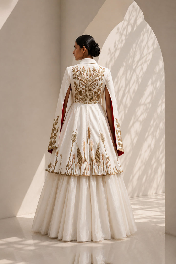 Ivory ensemble featuring intricate gold embroidery, dramatic cape sleeves, and layered volume for a look that is both regal and contemporary.