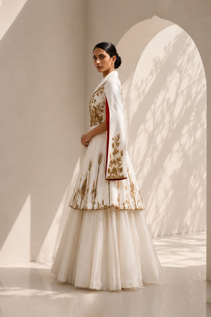Ivory ensemble featuring intricate gold embroidery, dramatic cape sleeves, and layered volume for a look that is both regal and contemporary.