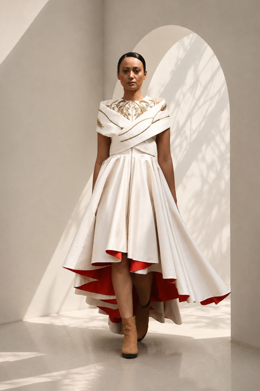 Ivory high-low dress featuring sculptural draped detailing, delicate embroidery, and a striking scarlet contrast lining for a bold yet refined statement.
