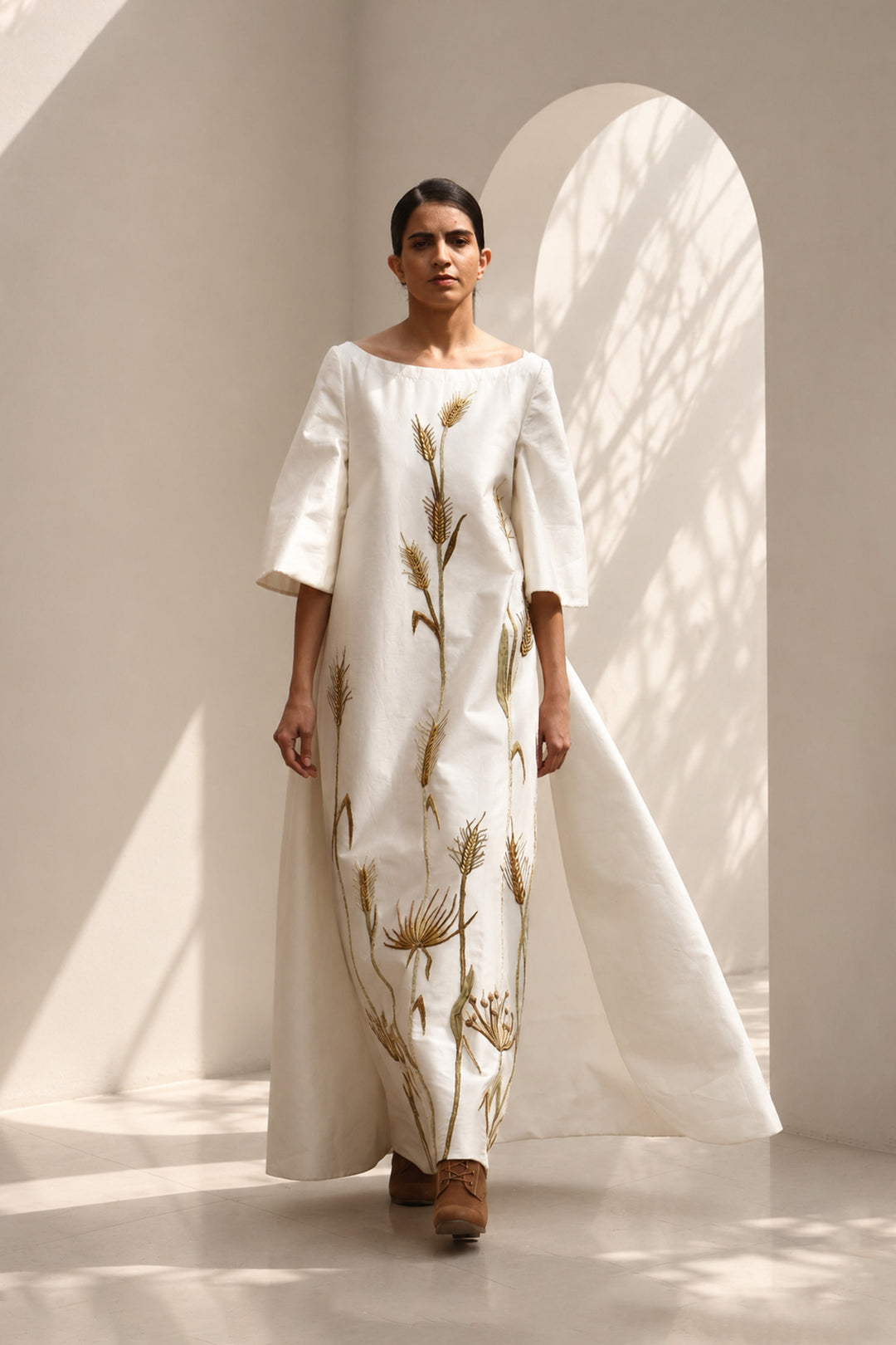 Ivory column gown adorned with delicate botanical embroidery. Featuring elegant boat-neckline, relaxed three-quarter sleeves &amp; flowing back panel.