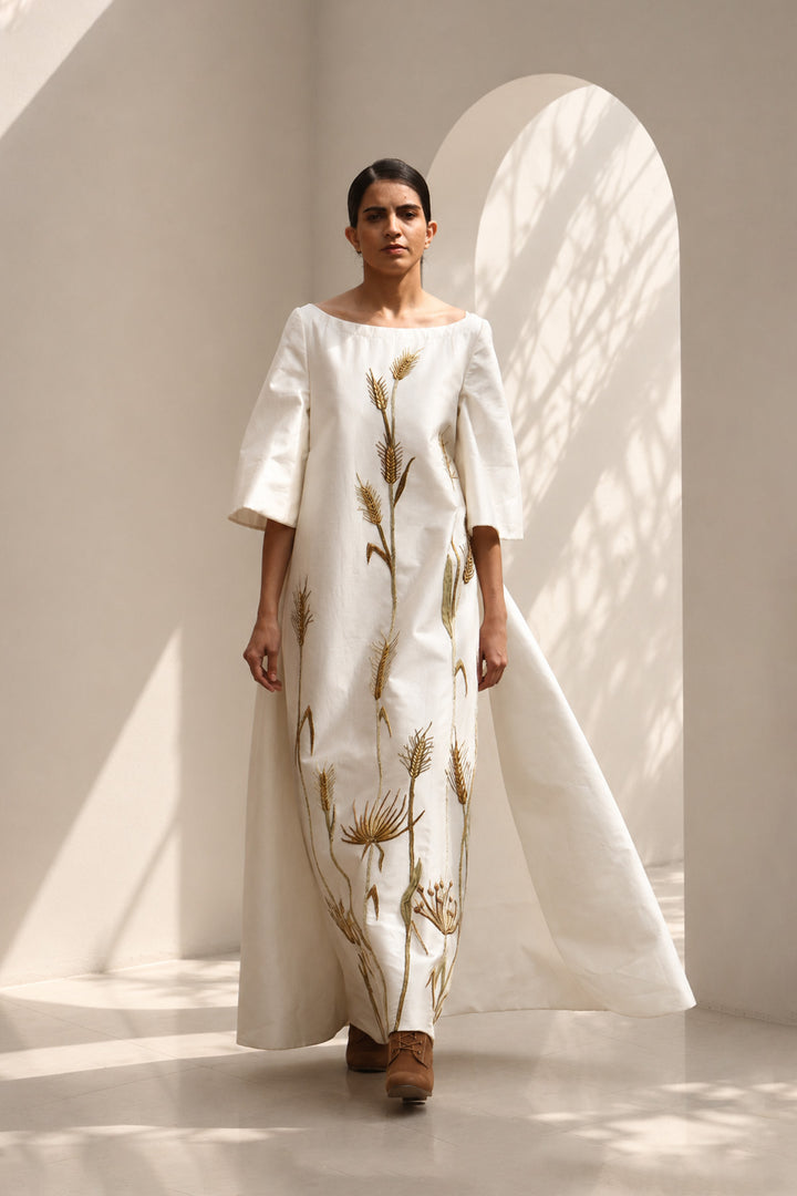Ivory column gown adorned with delicate botanical embroidery. Featuring elegant boat-neckline, relaxed three-quarter sleeves &amp; flowing back panel.