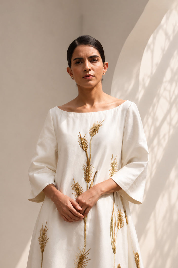Ivory column gown adorned with delicate botanical embroidery. Featuring elegant boat-neckline, relaxed three-quarter sleeves &amp; flowing back panel.